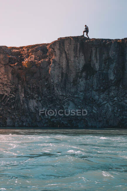 Side view of traveler walking on top of rocky cliff above blue waves in sunlight — Stock Photo
