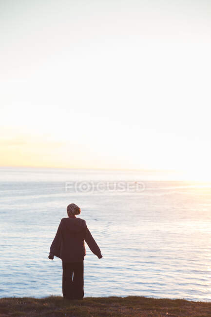 Rückansicht einer Frau, die auf einer Klippe steht, mit leuchtend blauem Meer im Hintergrund im Gegenlicht — Stockfoto