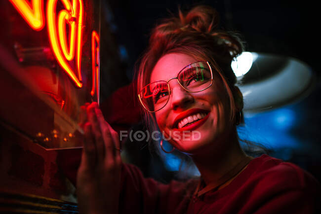 Vista lateral da mulher atraente em óculos com os olhos fechados inclinados na parede no sinal de néon de iluminação — Fotografia de Stock