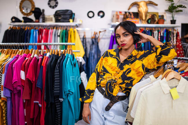 Bella giovane signora in abito elegante appoggiato su vestiti rail e guardando la fotocamera mentre in piedi in un piccolo negozio — Foto stock