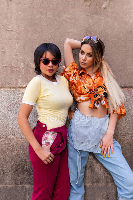 Two young ladies in trendy outfits leaning on building wall while standing on city street on sunny day — Stock Photo