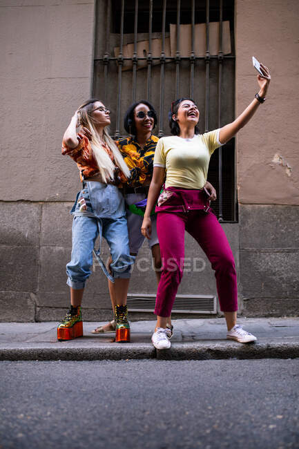Grupo de jóvenes en trajes de moda riendo y tomando selfie mientras se divierten en la calle de la ciudad - foto de stock