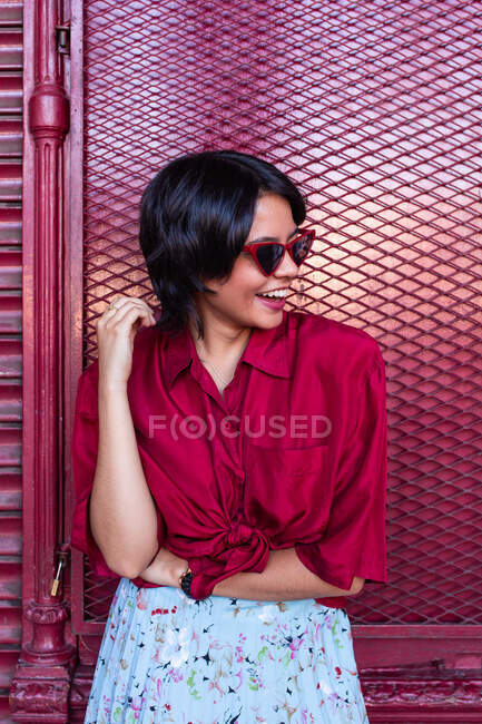 Красива молода жінка в модному вбранні сміється і дивиться геть, спираючись на бургундську стіну на міській вулиці — стокове фото