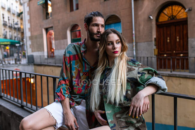 Young man and woman in trendy outfits sitting on metal fence on city street and looking at camera — Stock Photo