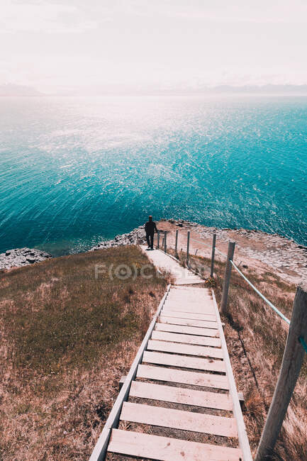 From above back view of tourist walking down wooden stairs towards beautiful rocky coastline with turquoise water in sunlight — Stock Photo