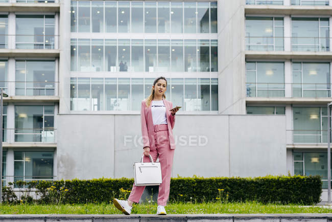 Стильная молодая женщина со смартфоном в розовом стоит перед современным офисным зданием — стоковое фото