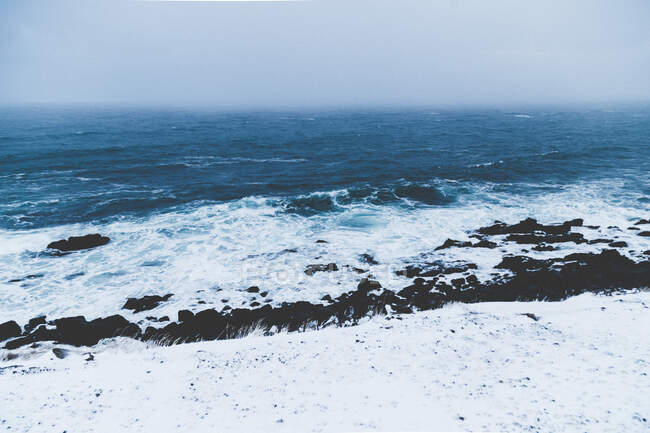 Landscape of cold ocean waves dashing against snowy rocky shoreline in mist — Stock Photo