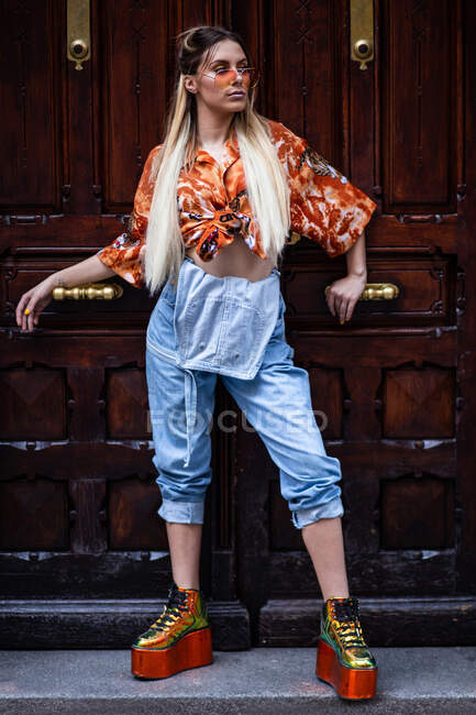 Bella giovane donna in abito alla moda guardando altrove mentre si appoggia sulla porta di legno di lusso sulla strada della città — Foto stock