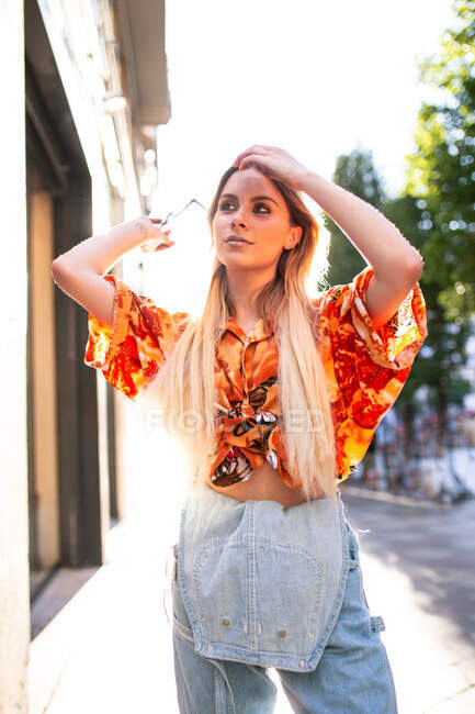 Bela jovem fêmea na roupa da moda ajustando o cabelo e olhando para cima, enquanto de pé na rua da cidade no dia ensolarado — Fotografia de Stock