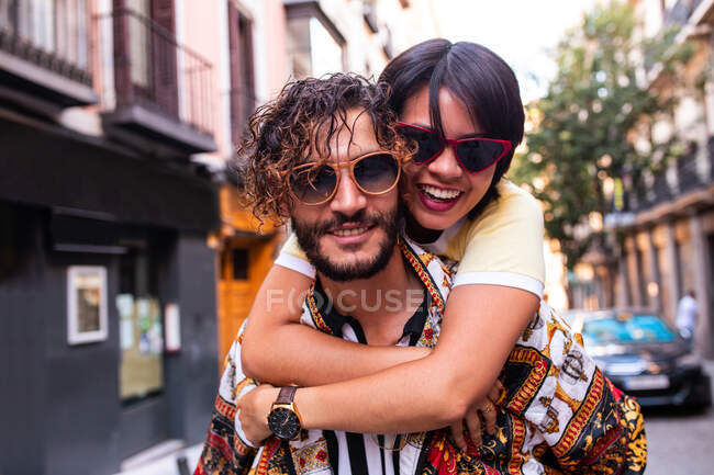Handsome bearded man in trendy outfit smiling and giving piggyback ride to cheerful girlfriend on blurred background of city street — Stock Photo