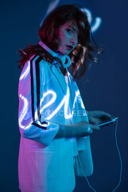 Женщина слушает музыку и использует смартфон в неоновом свете надпись и смотреть на камеру — стоковое фото