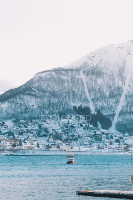 Вид катера в голубой воде на фоне маленького городка на берегу со снежной горой над головой — стоковое фото