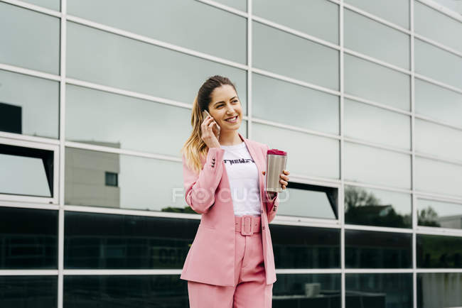 Cheerful stylish woman with coffee talking on smartphone in front of modern office building — Stock Photo