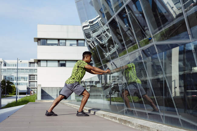 Vista laterale dell'uomo etnico adulto che si estende al corrimano in vetro mentre si allena sulla moderna strada della città — Foto stock