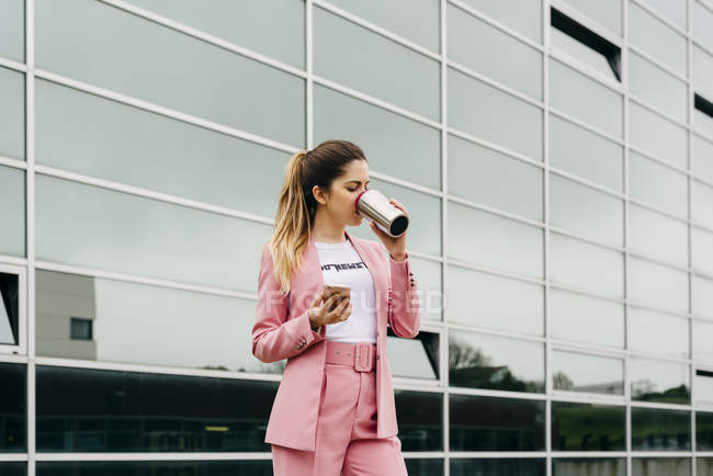Trendy woman drinking coffee while standing in front of modern office building — Stock Photo