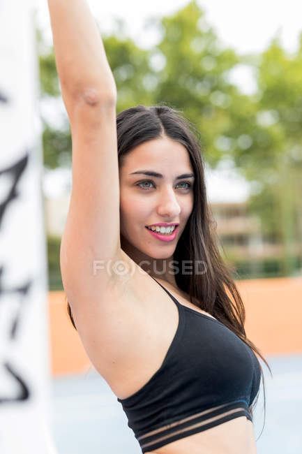 Молодая женщина в спортивном топе стоит на открытом воздухе с поднятой рукой и смотрит в сторону — Вертикаль, брюнетка - Stock Photo