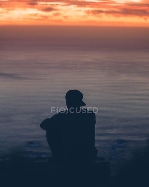 Rückansicht einer unkenntlichen Person, die auf einem Felsen sitzt und den Sonnenuntergang am Meer bewundert. — Stockfoto