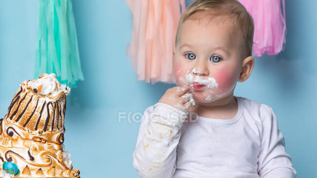 Lindo niño sentado en el pastel de cumpleaños y comerlo con la mano sobre fondo azul. - foto de stock