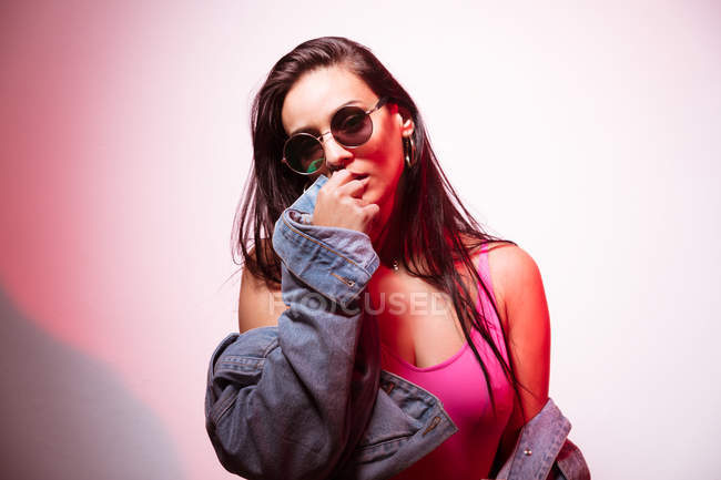 Красивая молодая розовая женщина в солнечных очках стоит у белой стены и смотрит в камеру — стоковое фото