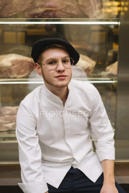 Junger Mann in eleganter weißer Jacke und Mütze mit modischer Brille und Blick in die Kamera. — Stockfoto