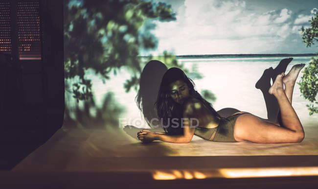 Young sensual woman in one piece swimsuit lying in studio on background of beach — Stock Photo