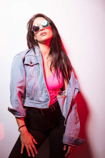 Молода рожева одягнена жінка в сонцезахисних окулярах стоїть на білій стіні і дивиться на камеру — стокове фото