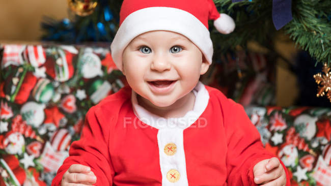 Portrait of happy little boy in Santa Claus costume sitting at Christmas tree — Stock Photo