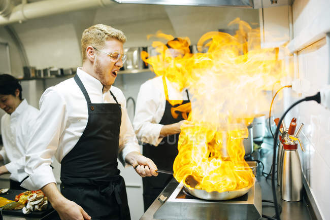 Cozinheiro feliz fazendo um flambe na cozinha do restaurante com colega assistindo — Fotografia de Stock