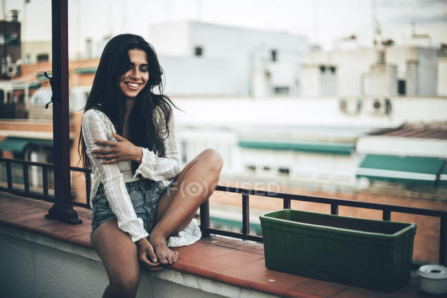 Молода щаслива жінка сидить на балконі і посміхається на тлі будинків — стокове фото