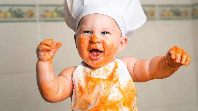 Niño alegre en delantal con la cara sucia cubierta de salsa. - foto de stock