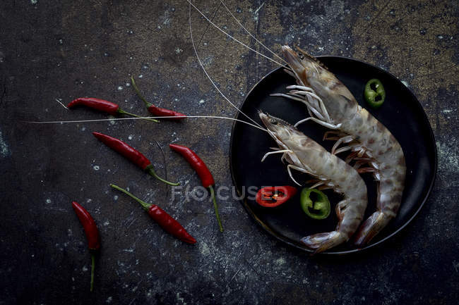 Raw tiger prawns with small red peppers on plate on dark tabletop — Stock Photo