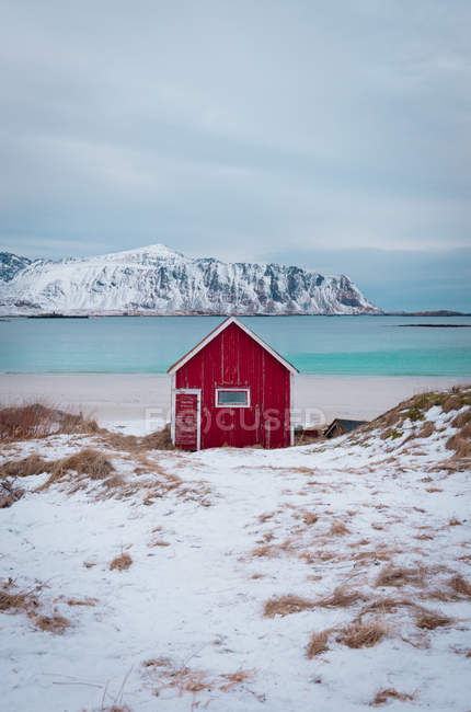 Pequena cabine de madeira vermelha no litoral nevado com água do mar azul e montanhas no fundo, Lofoten, Noruega — Fotografia de Stock