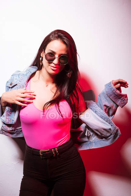 Seducing young pink dressed woman in sunglasses standing at white wall with hand in hair — Stock Photo