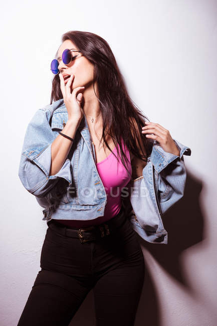 Séduire jeune femme rose habillée en lunettes de soleil debout au mur blanc — Photo de stock