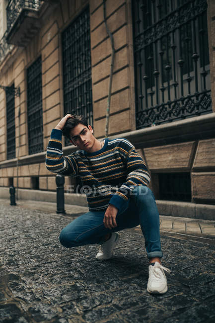 Молодой стильный подросток в свитере приседает на городской улице и смотрит в камеру — стоковое фото