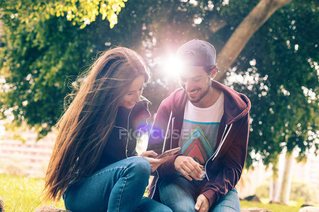 Lachendes junges Paar sitzt mit Smartphone im Park — Stockfoto