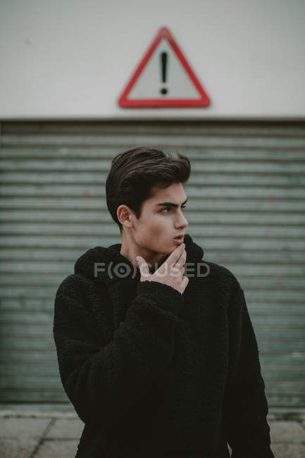Thoughtful teenager in black hooded jacket standing on street with exclamation sign and looking away — Stock Photo