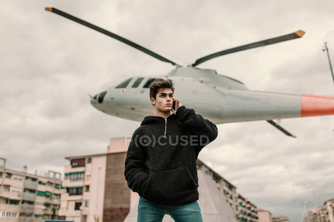 Bonito jovem de pé no monumento de helicóptero na cidade e falando no smartphone — Fotografia de Stock