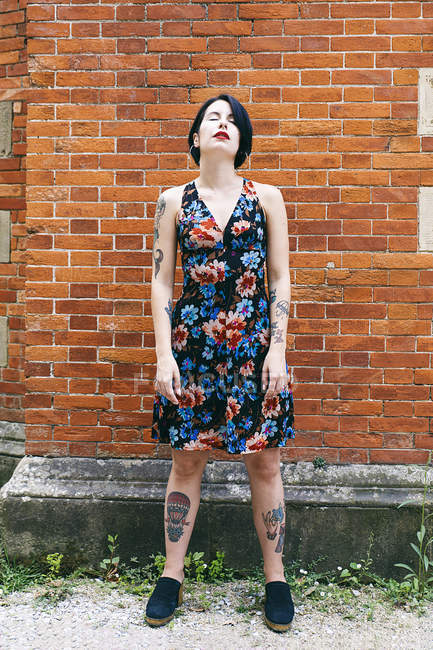 Татуйована жінка в візерунковому платті, що стоїть перед цегляною стіною — стокове фото