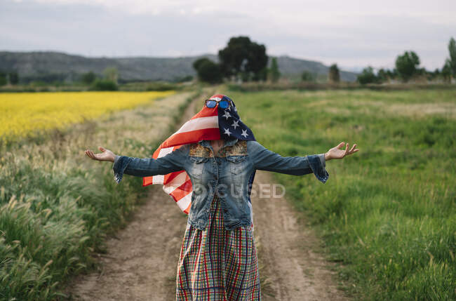 Senhora em roupas casuais ficar em campo amarelo com óculos de sol sobre bandeira americana com luz solar — Fotografia de Stock