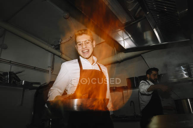 Cocinero haciendo un flambe en la cocina del restaurante con colega en el fondo - foto de stock