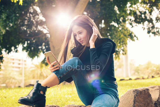 Jeune femme assise sur le rocher et utilisant un smartphone dans le parc — Photo de stock