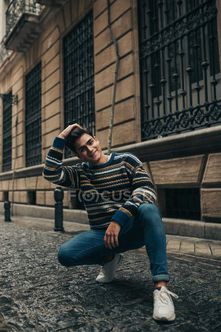 Joven sonriente adolescente con estilo en suéter agacharse en la calle de la ciudad y mirando a la cámara - foto de stock