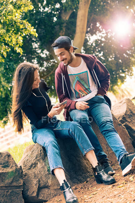 Sonriente joven pareja sentada en roca con smartphone en el parque - foto de stock