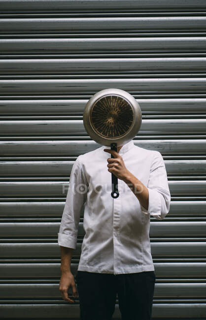 Pensive young man in glasses and white jacket covering face with frying pan looking up. — Stock Photo