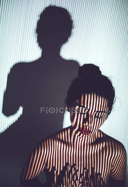 Naked young woman covering breast with hands in studio with red lips and striped shadow on face and body — Stock Photo