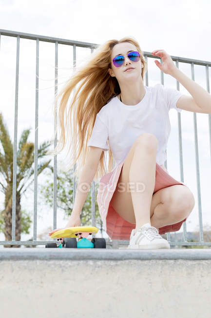 Blonde fille élégante dans des lunettes de soleil assis dans un skate park avec penny board — Photo de stock