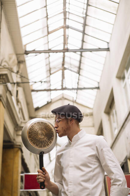 Stylish romantic man in chef jacket and black cap kissing frying pan devoted to culinary and cooking. — Stock Photo