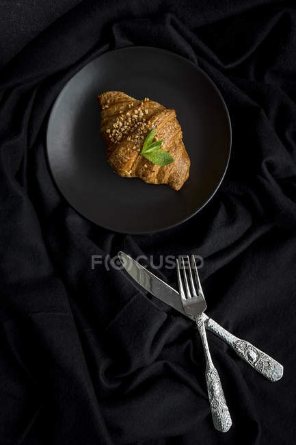 Baked croissant on plate on black fabric with knife and fork — Stock Photo