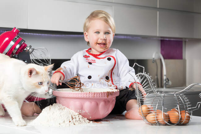 Ragazzino seduto su un tavolo disordinato con gatto e giocare con gli ingredienti per cucinare. — Foto stock